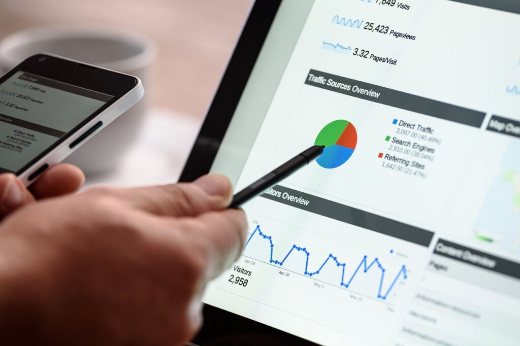 An individual is holding a smartphone and pointing at a tablet screen displaying various graphs and charts. The screen shows data on website visitors, traffic sources, and analytics with a pie chart, line graph, and numerical figures. This arrangement ensures comprehensive security by monitoring all hosting metrics in real-time.