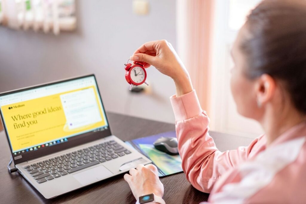 Woman working at a laptop, holding a red alarm clock.
